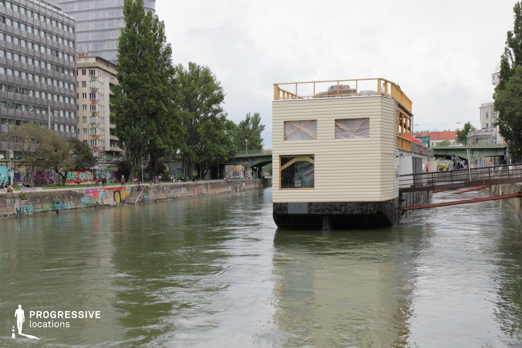 This setting presents an industrial waterway scene with a floating structure docked along a graffiti-covered concrete embankment. The structure, featuring beige siding and large windows, has an unfinished appearance, adding an element of urban rawness. Overhead, a green metal bridge spans the water, connecting the banks, while modern office buildings and dense greenery frame the background. The juxtaposition of natural and industrial elements creates a visually rich contrast, making this location ideal for filming contemporary urban dramas, independent films, or gritty crime thrillers. The reflective water surface and layered architectural details enhance its cinematic depth.