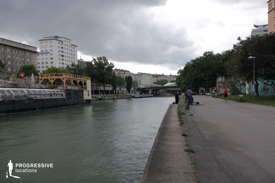 This riverside location presents a mix of natural and industrial elements, featuring a long paved walkway parallel to the water, lined with trees and graffiti-covered walls. The opposite bank showcases a variety of architectural styles, from mid-century buildings to modern structures, along with a floating venue with a wooden terrace, adding an urban waterfront vibe. The river itself provides a strong linear perspective, leading toward bridges and distant city structures. This versatile setting is ideal for filming introspective, dramatic, or urban crime scenes