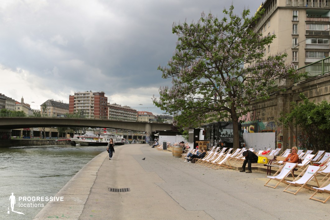 This urban riverside promenade features a wide, paved walkway running parallel to the water, flanked by a row of wooden lounge chairs on a raised platform. A large, blossoming tree adds a natural touch, while a food truck and graffiti-covered stone walls contribute to the setting’s modern, edgy character. The adjacent river is crossed by a concrete bridge, with mid-rise residential and commercial buildings forming the backdrop. The location’s mix of industrial and relaxed elements makes it suitable for filming urban lifestyle sequences, casual social interactions, or atmospheric cityscape montages. Ideal for contemporary dramas, travel documentaries, or indie films focusing on urban culture.