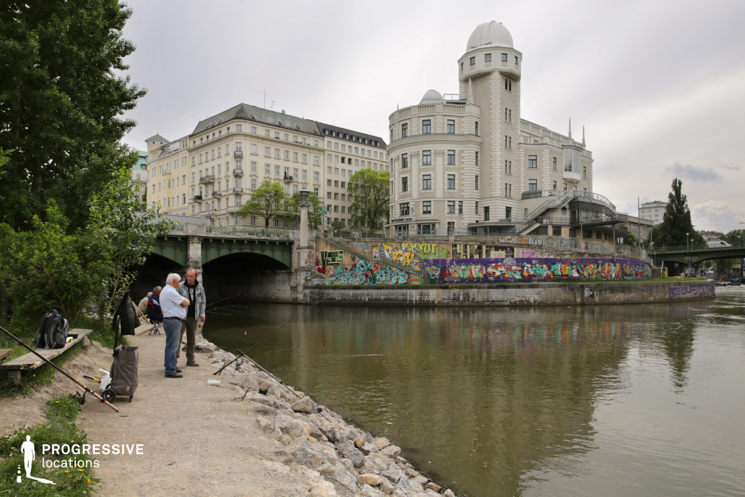 This riverside location blends classic and modern architectural elements, featuring an imposing historic building with a distinctive dome and intricate façade, alongside a green metal bridge that connects two urban areas. The riverbank is lined with graffiti-covered walls, adding a raw, contemporary contrast to the refined buildings. A rocky shoreline and a dirt path create a natural foreground, leading toward the water, enhancing the depth of the composition. This setting is well-suited for crime dramas, urban documentaries, and reflective or atmospheric scenes, offering a mix of historical elegance and gritty street culture.