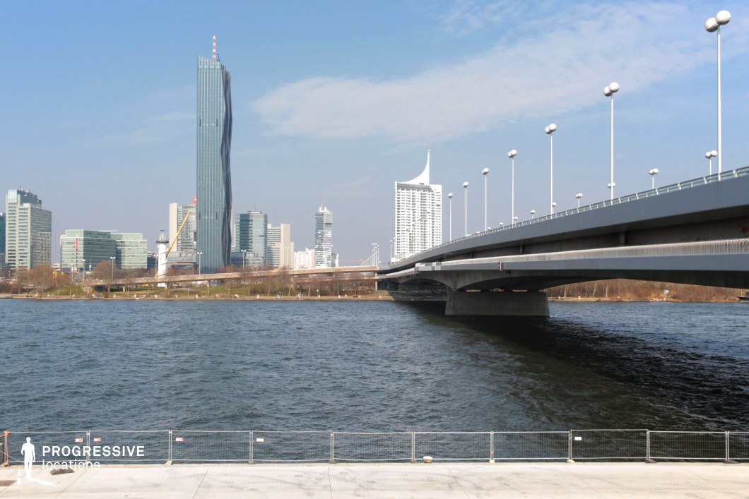 This striking urban scene features a sleek modern bridge stretching across a wide river, leading towards a skyline of contemporary high-rises. The most prominent structure is a twisting glass skyscraper, contrasting with the geometric forms of the surrounding buildings. The bridge is lined with evenly spaced, spherical streetlights, adding a sense of symmetry and scale. The riverbank in the foreground provides a strong visual anchor, enhancing the depth and openness of the composition. This location is ideal for filming futuristic cityscapes, action sequences, corporate dramas, or espionage thrillers, offering a setting that conveys modernity, movement, and urban sophistication.