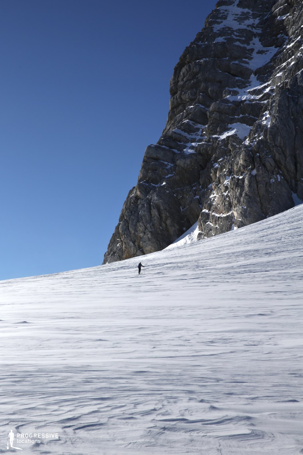This natural location presents a vast, snow-covered slope dominated by a towering vertical rock face rising sharply on the right side. The rugged, dark grey stone surface features pronounced stratification and jagged textures, offering strong contrast against the smooth, wind-sculpted patterns of the snowfield below. The terrain gently inclines upward, while the camera angle—shot from a low perspective—emphasizes the scale and verticality of the landscape. The open composition and stark geological formations create a visually striking backdrop. This setting is ideal for film scenes requiring isolation, raw natural elements, or dramatic tension, making it especially suitable for survival, adventure, thriller, or documentary-style productions.