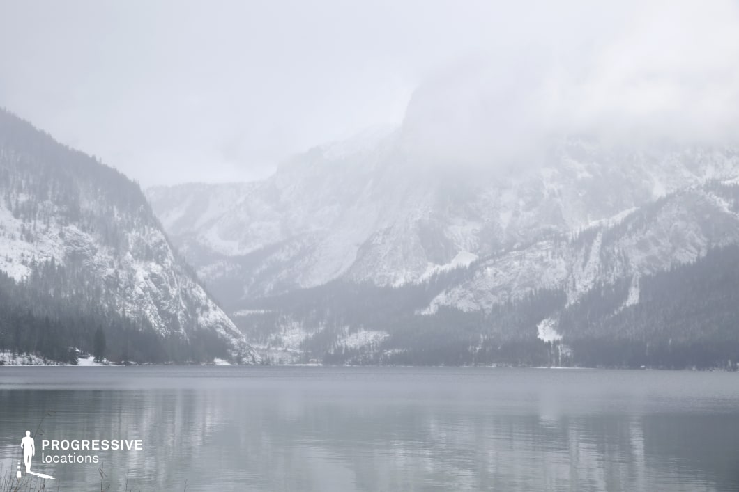 This expansive natural location features a wide, still lake bordered by snow-covered forested mountains and steep rock faces. The visual composition is dominated by cool, desaturated tones—mainly whites, greys, and muted greens—creating a soft, atmospheric aesthetic. The mountains display a strong vertical scale and textural contrast, with jagged stone formations emerging beneath the tree lines. The reflective water surface enhances depth and symmetry in the frame, while the distant mountain ridges gradually fade into a misty backdrop, generating a layered spatial effect. The absence of modern infrastructure and the vast openness of the landscape emphasize a remote and untouched quality. This location is ideal for films requiring serene, introspective, or isolating environments, and lends itself well to genres such as drama, mystery, or historical epics, particularly for scenes set in remote wilderness or involving natural grandeur.