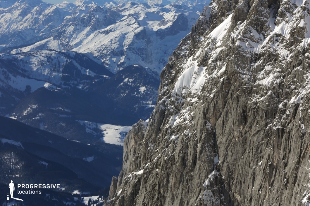 This dramatic mountainous location showcases a steep, jagged rock face partially covered with snow, sharply contrasting with the layered, forested slopes and distant snow-capped ranges in the background. The cliff itself consists of vertically striated grey stone with deep crevices and an angular surface geometry, emphasizing its raw and imposing character. The backdrop of undulating valleys and ridges provides significant visual depth and scale, while the clear delineation between the cliff in the foreground and the expansive mountain landscape behind it enhances the spatial dynamics of the composition. The location’s striking natural formation, varied elevation, and unspoiled terrain make it especially suitable for filming intense, adventurous, or contemplative sequences. It is ideal for genres such as drama, thriller, and action, particularly for establishing shots, wide landscape sequences, or scenes that require a sense of isolation or grandeur.