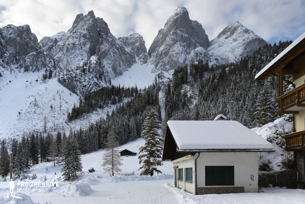 This location presents a dramatic alpine valley framed by steep, snow-covered rocky peaks and dense coniferous forest that ascends sharply toward the rugged mountains. In the foreground, a modest white building with a low-pitched roof and shuttered windows introduces a rural architectural element, while additional wooden structures with visible balconies suggest a blend of traditional alpine style. The snow-blanketed landscape emphasizes clean lines and high contrast, enhancing the sharp silhouettes of the pine trees and the jagged mountain ridges. The perspective offers a deep compositional axis, moving from the flat, accessible foreground into the vertical drama of the rocky elevations. The combination of natural monumentality and structured habitation creates a visually balanced and textured scene. This location is ideal for filming dramatic, nature-driven narratives or contemplative sequences, particularly in genres such as adventure, drama, or mystery, and lends itself well to both wide establishing shots and detailed close-ups that explore the relationship between built environment and untamed wilderness.