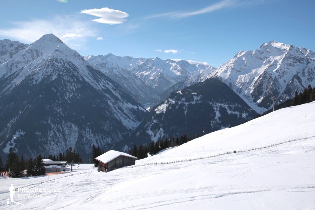 This location captures a panoramic alpine landscape dominated by steep, snow-covered mountain ridges stretching across the horizon. In the foreground, a gently sloping hillside is blanketed in pristine snow, interrupted only by a solitary wooden cabin and subtle fencing lines that follow the terrain’s curvature. The central valley between the mountains creates a strong visual axis, emphasizing depth and scale. The dominant colors are crisp whites and deep forest greens, contributing to a stark yet harmonious contrast that defines the winter environment. The rugged peaks exhibit dramatic elevation changes and natural symmetry, while the hillside maintains a smooth, flowing geometry ideal for wide-angle cinematic shots. This location is particularly suitable for filming scenes requiring expansive natural backdrops, offering visual clarity and structural simplicity, making it ideal for drama, adventure, or nature-themed productions.