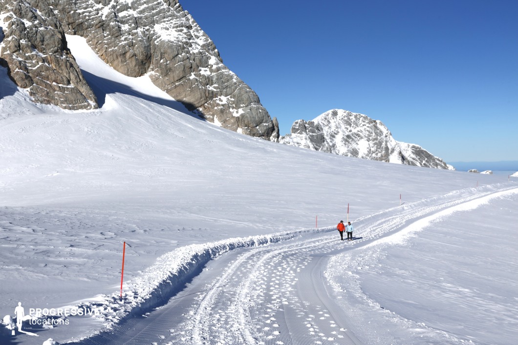 This high-altitude location features a broad, winding snow-covered path that leads through a pristine alpine landscape, framed by steep, rugged rock formations partially covered with compacted snow. The terrain is open and expansive, offering a dramatic contrast between the white snowfield and the exposed gray stone faces of the surrounding peaks. The surface of the pathway is groomed with visible parallel texture lines, creating a sense of order within the natural environment, while the gentle curves of the path introduce a subtle geometric rhythm. The vertical rock walls and distant ridges establish a strong visual backdrop, enhancing the location’s sense of scale and isolation. Ideal for filming scenes requiring remote, extreme or majestic settings, this location is particularly suited for genres such as adventure, survival, fantasy, or drama, and works well for wide establishing shots or movement-based sequences like treks or ascents.