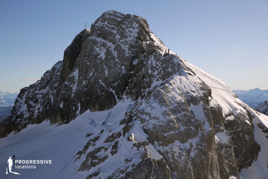 This dramatic mountain landscape showcases a sharply contoured alpine ridge, characterized by steep, jagged rock formations partially covered with snow. The dominant hues are cold greys and whites, which enhance the stark, imposing presence of the rock face against the clear backdrop. The rugged rock texture combined with the smooth, sweeping snowfields creates a visually dynamic composition with strong vertical and diagonal lines. The terrain includes a narrow path or ridge traverse with occasional structural elements integrated into the mountain, hinting at its accessibility for alpine infrastructure or cableway stations. This elevated and visually commanding setting is ideal for cinematic use, particularly in adventure, thriller, or dramatic genres. Its isolated, awe-inspiring scale and natural grandeur make it well-suited for establishing shots, high-stakes sequences, or scenes requiring a sense of remoteness and intensity.