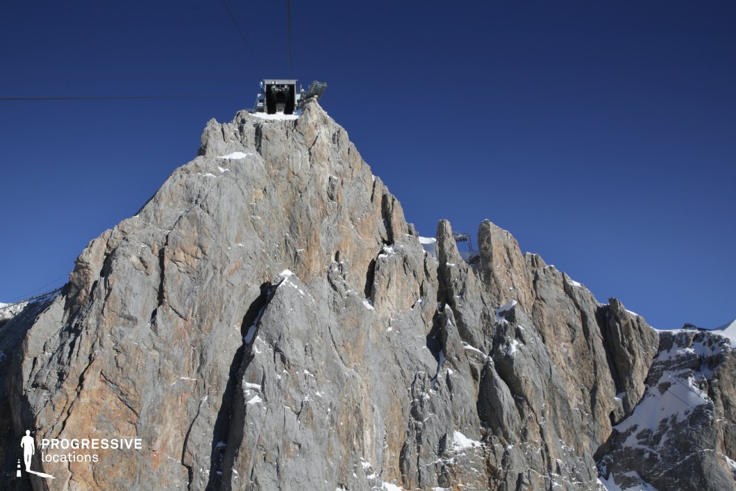 This dramatic mountain peak features a rugged, vertical rock formation composed of sharp, jagged edges in varying shades of grey and ochre, accented by scattered patches of snow. At the summit, a prominent cable car station emerges as a striking man-made structure contrasting against the raw geological landscape. The bold geometry of the rocks, paired with the engineered lines of the cable car wires, creates a visually arresting composition. The texture of the stone is coarse and fractured, emphasizing the natural severity of the high-altitude environment. The perspective, taken from a low vantage point looking upward, reinforces the towering scale of the mountain and the isolation of the station perched at its pinnacle. This location is ideal for action, thriller, or adventure genres, particularly for scenes requiring an extreme alpine environment, a sense of isolation, or a high-stakes setting with visual grandeur.