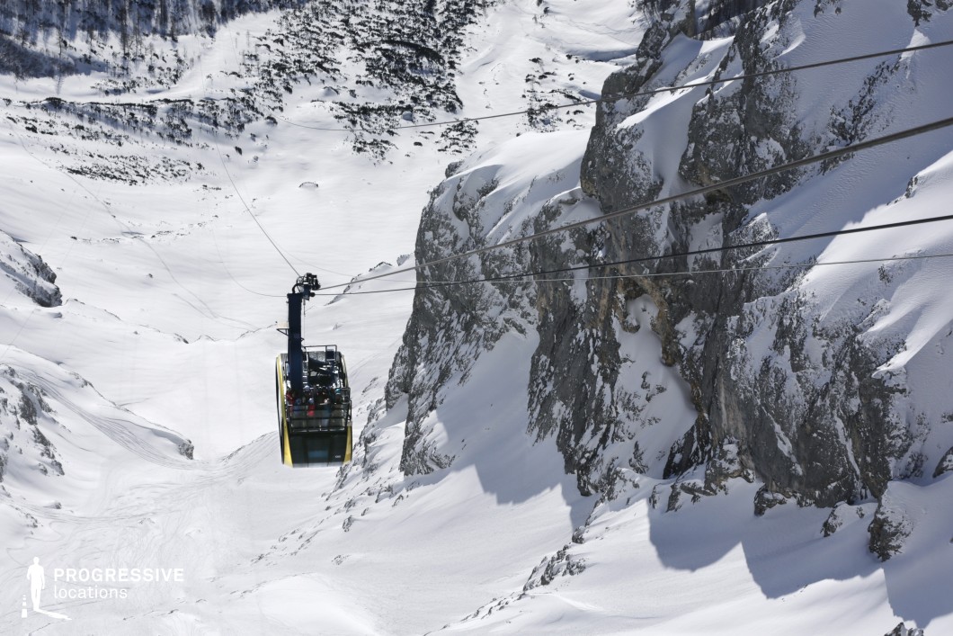 This high-altitude location features a dramatic aerial cable car suspended over a steep, rugged mountain landscape. The terrain is dominated by angular, snow-covered rock faces and expansive slopes that stretch across the scene, providing strong linear and geometric elements. The contrast between the sheer vertical cliff and the horizontal cables intensifies the location’s dramatic verticality. The materiality of the scene includes raw, jagged stone and deep, uninterrupted snow cover, contributing to a stark and monumental atmosphere. The perspective is slightly elevated, capturing the cable car mid-air as it traverses between ridgelines, enhancing the sense of isolation and vastness. This location is ideal for filming scenes that require a remote, imposing natural setting, particularly in thriller, adventure, or documentary genres, and is well-suited for sequences emphasizing altitude, transition, or environmental extremes.