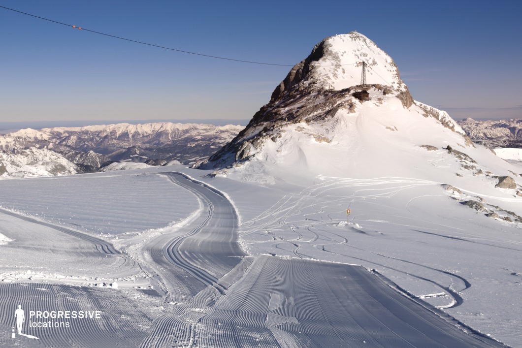 This high-altitude mountain landscape features a vast snow-covered plateau dominated by a distinct rocky peak, partially covered in snow and equipped with a functional lift infrastructure. The foreground showcases meticulously groomed ski tracks with symmetrical, linear textures that emphasize the terrain’s man-made precision and expansive width. The snow appears densely packed and evenly layered, contrasting with the rougher, untouched slopes on the right side. In the background, a panoramic ridge of distant mountain ranges creates a visually continuous and immersive horizon line. The open composition, marked by clear spatial layering and strong geometric lines, makes this location ideal for cinematic wide shots, aerial sequences, and scenes emphasizing isolation or scale. Suitable for action, thriller, or documentary genres, this site offers a dynamic yet controlled natural environment.
