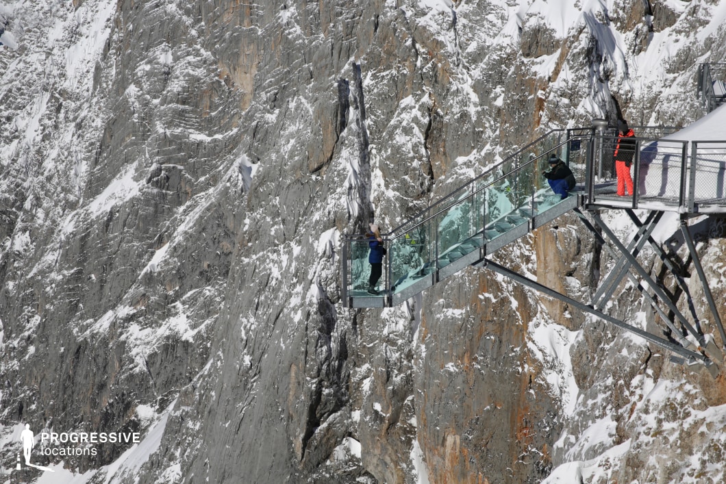 This dramatic location features a narrow glass-bottomed skywalk cantilevered from a steep, snow-dusted rock face. The platform, constructed from steel supports and transparent reinforced glass, extends outward into open space, offering an unobstructed, vertigo-inducing perspective over a rugged alpine landscape. The surrounding mountain surface is composed of jagged grey stone with sharp vertical striations and patches of snow, creating a stark natural backdrop. The materials—glass, steel, and raw rock—form a striking contrast between modern engineering and untouched geology. The setting is both visually intense and architecturally minimal, with high-altitude exposure enhancing its dramatic appeal. This location is ideal for filming scenes that require an extreme or awe-inspiring natural environment, such as thrillers, documentaries, or high-concept fashion shoots where isolation, elevation, and panoramic depth are essential visual elements.