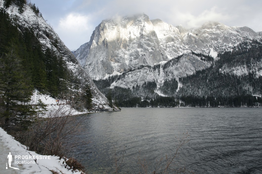 This location presents a striking natural landscape defined by a tranquil alpine lake bordered by steep, forested slopes and dominated by a dramatic, snow-covered mountain backdrop. The terrain transitions from densely wooded hillsides with evergreen trees to towering rock faces that exhibit sharp verticality and rugged textures. The dominant color palette is composed of muted greys, deep greens, and soft whites, producing a stark yet harmonious contrast that enhances the location’s visual depth. The composition is balanced, with the expansive water body in the foreground offering a reflective surface that contrasts with the imposing geological formations. The image is taken from a slightly elevated side angle, allowing both depth and perspective, and emphasizing the spatial scale between lake, forest, and mountain. This location is highly suitable for productions requiring a remote, raw natural environment—ideal for nature documentaries, dramatic narratives, or introspective scenes in adventure, fantasy, or survival genres.