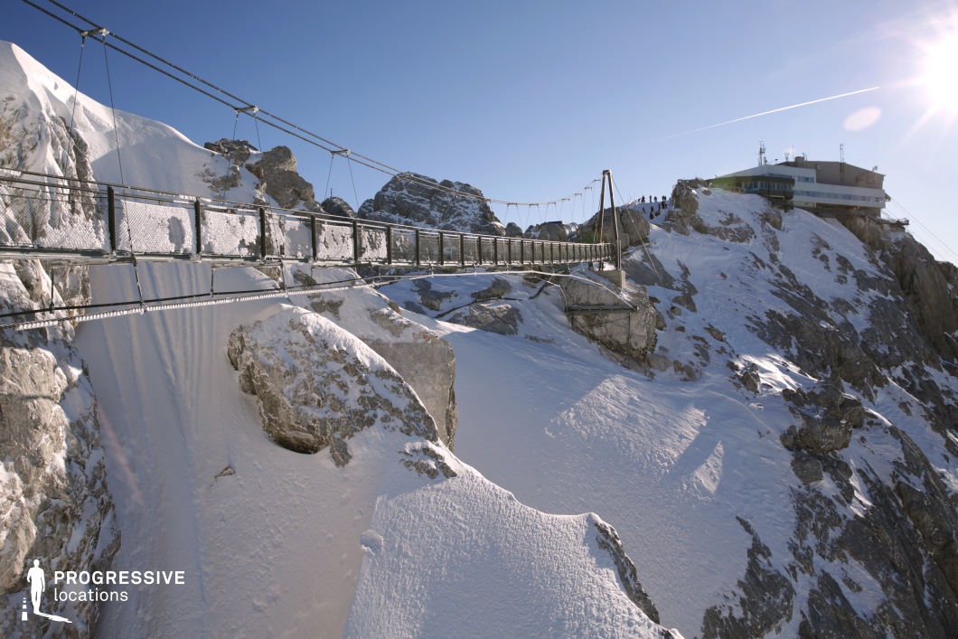 This striking mountain location features a steel suspension bridge spanning a steep, snow-covered ravine between two rugged rock formations. The bridge's sturdy metal frame and mesh safety railings contrast with the surrounding natural stone and snow textures, highlighting the harsh and remote alpine setting. Dominated by shades of white and gray with muted steel tones, the visual composition is clean, stark, and dramatic. To the right, a contemporary mountain structure with a low, horizontal profile is integrated into the rocky summit, enhancing the location’s functional realism. The elevated perspective and expansive background reinforce the location’s vertical scale and sense of isolation. This visually commanding and extreme setting is highly suitable for action, thriller, or expedition-themed productions, particularly for scenes requiring tension, altitude, or raw natural elements.
