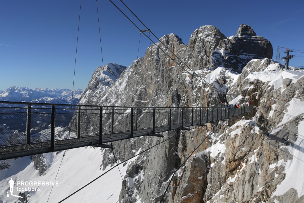 This dramatic mountain location features a narrow suspension bridge stretching across a deep alpine chasm, anchored between rugged, snow-covered rock faces. The steel structure is composed of metal mesh panels and tensile cables, offering both transparency and stability, and establishing a modern, industrial contrast against the raw natural backdrop. The surrounding mountains exhibit sheer vertical cliffs, jagged peaks, and stratified rock surfaces, with dominant hues of grey and white that evoke a stark, high-altitude environment. The visual depth is enhanced by the extreme elevation and vast panoramic vista of distant mountain ranges. With its bold geometric lines and open-air exposure, this location is ideal for high-stakes, physically intense, or psychologically charged scenes. It is particularly suited for action, adventure, thriller, or documentary-style productions requiring a visually imposing and isolated alpine setting.