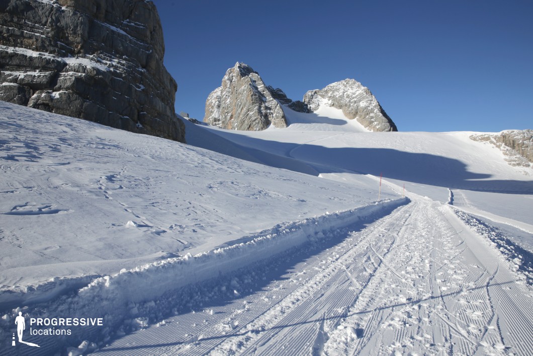 This image captures a wide, open mountain landscape defined by a meticulously groomed snow-covered path that cuts through a vast alpine expanse. The dominant visual elements include rugged, stratified rock formations and smooth, sweeping snowfields that create dramatic contrasts in texture and elevation. The monochromatic palette, composed primarily of whites and cold grays with hints of stone beige, establishes a clean, minimalist aesthetic. The jagged cliffs and towering peaks in the background provide a strong geometric focal point, while the subtle curvature of the path adds depth and directional flow. The composition is visually expansive, allowing for strong horizon lines and long-range perspectives ideal for cinematic framing. This location is ideal for filming scenes requiring remote, high-altitude, or survivalist settings, and suits genres such as drama, adventure, or thriller, especially where isolation and vast natural scale are thematically relevant.