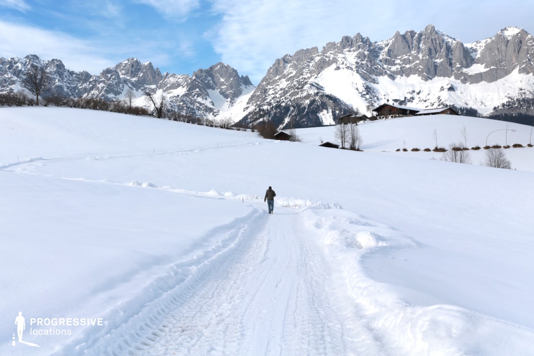 This location presents a wide, snow-covered rural pathway that gently inclines toward a series of small alpine buildings nestled at the base of a dramatic mountain range. The path is clearly defined by compacted snow and tire tracks, adding a textural contrast to the otherwise smooth, untouched snowfields on either side. Towering, jagged peaks dominate the background, their grey rock faces partially veiled by layers of snow, creating a strong geometric presence against the open sky. The wooden buildings scattered along the hillside are low and rustic in style, integrating naturally into the landscape and reinforcing a sense of remote, high-altitude simplicity. The setting is visually expansive and offers a deep perspective from the foreground path to the distant mountains, making it an excellent location for filming wide establishing shots, introspective travel sequences, or scenes requiring a remote and rugged environment. It is particularly suitable for drama, nature documentaries, or adventure genres.