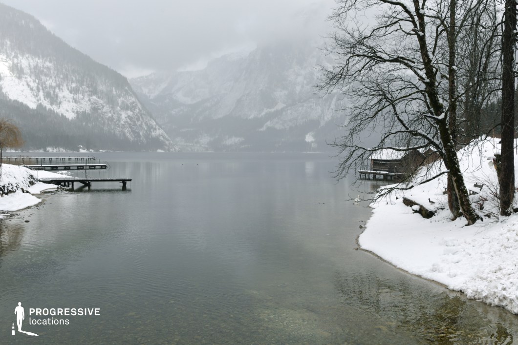 This natural lakeside setting features calm, clear water bordered by a snow-covered shoreline and steep, forested mountains in the background. On both sides of the image, wooden docks and piers extend into the lake, adding linear geometric contrast to the organic shoreline and bare trees. The presence of traditional wooden structures built on stilts over the water introduces a rustic architectural element that complements the serene, remote ambiance. The perspective is taken from a slightly elevated frontal view, with a strong sense of depth emphasized by the layering of foreground trees, middle-ground water, and distant mountainous slopes. The consistent muted color palette of whites, greys, and dark browns reinforces a minimalist, tranquil atmosphere. This location is highly versatile for filming, lending itself well to genres such as drama, psychological thrillers, or art films, particularly for scenes requiring a quiet, introspective, or isolated natural backdrop.