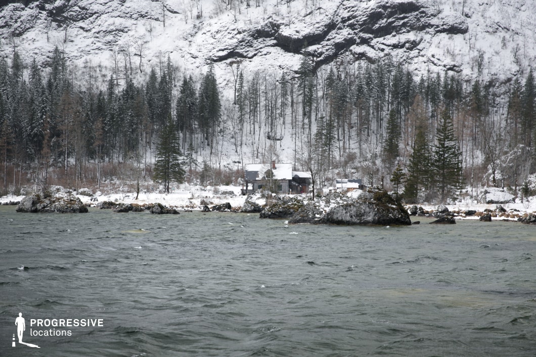 This remote location captures a rugged lakeside scene framed by a steep, snow-covered forested hillside and scattered boulders along the shoreline. In the distance, a weathered wooden cabin complex sits discreetly at the edge of the pine and deciduous tree line, its muted tones blending naturally into the subdued winter palette of grays, whites, and deep greens. The uneven shoreline is punctuated by large rocks that protrude into the water, enhancing the wild, untouched character of the area. The composition emphasizes horizontal layering—from the textured surface of the water in the foreground, to the treeline and vertical cliff face rising in the background. This site is particularly well-suited for genres such as psychological thrillers, survival dramas, or mystery films, offering a visually striking yet isolated setting for scenes requiring seclusion, tension, or introspective atmosphere.