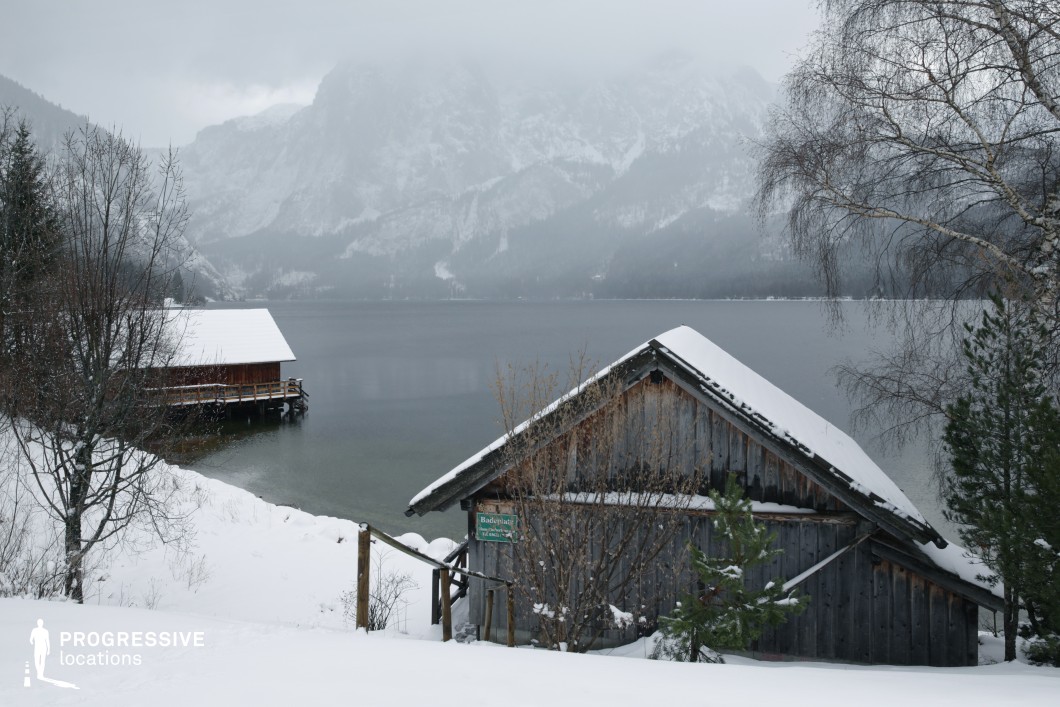 This location presents a pair of rustic wooden structures situated on the edge of a large, calm lake, set against a dramatic mountainous backdrop partially veiled by mist. The buildings, with their pitched roofs blanketed in snow, emphasize a traditional, unembellished architectural style that blends naturally with the alpine surroundings. The muted tones of aged timber and white snow dominate the visual palette, creating a quiet, atmospheric composition. The gentle slope leading to the water and the minimalistic fencing structure support an open spatial layout, while sparse vegetation and slender tree trunks introduce vertical visual elements. The elevated vantage point offers a broad perspective, enhancing depth and balance. This site is particularly suitable for introspective or atmospheric film genres such as drama or mystery, and can effectively support scenes requiring remote, nature-bound, or emotionally charged settings.