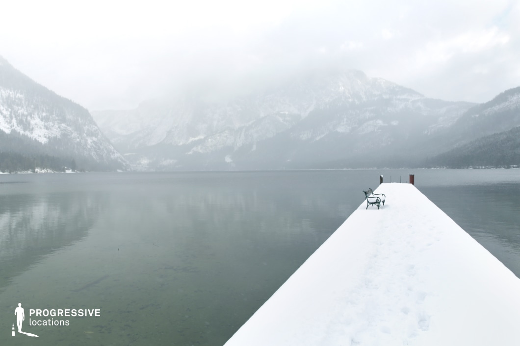 This location features a long, snow-covered dock extending into a glassy, calm lake, framed by towering forested mountains partially obscured by mist. The dock is geometrically strong, forming a sharp, linear perspective that draws the viewer’s gaze toward the horizon, enhancing the scene’s depth. The bench placed on the dock introduces a solitary, fixed structural element, adding visual anchor without clutter. The surrounding mountainous terrain is steep and imposing, with layers of ridges creating a dramatic natural amphitheater around the water. Dominated by shades of white, grey, and muted green, the color palette is subdued and uniform, contributing to a serene, introspective mood. This location is ideal for filming reflective, atmospheric, or emotionally driven scenes, and it suits genres such as drama, mystery, or psychological thrillers where a quiet, isolated, and immersive natural setting is essential.