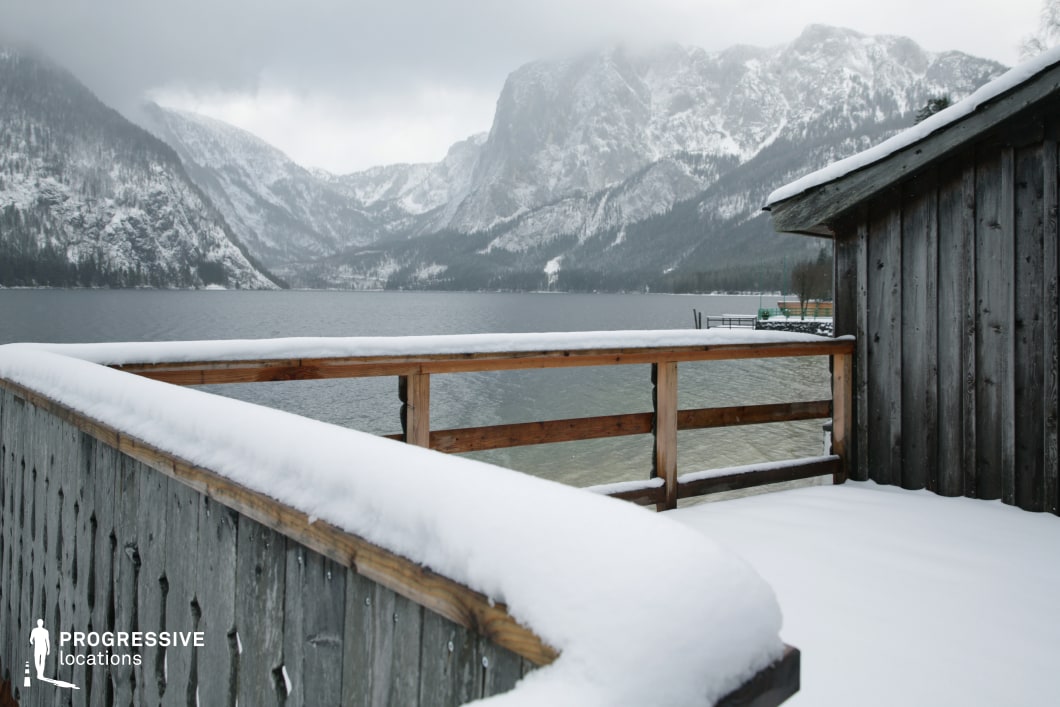 This lakeside terrace features a snow-covered wooden deck bordered by natural timber railings and weathered vertical plank siding, creating a rustic yet stable visual frame. The structure opens onto a calm lake, backed by towering, snow-dusted mountain formations that dominate the horizon in layered gradients. The viewpoint emphasizes the angular geometry of the deck, offering a semi-enclosed foreground that contrasts effectively with the expansive natural landscape beyond. The subdued color palette of muted greys, browns, and whites reinforces a grounded, contemplative mood. The overall spatial layout is compact but opens up dramatically toward the background, making it ideal for shoots requiring a contrast between intimate built structures and vast natural surroundings. Suitable for drama, arthouse cinema, or documentary-style productions, this location supports scenes requiring reflection, isolation, or quiet dialogue.