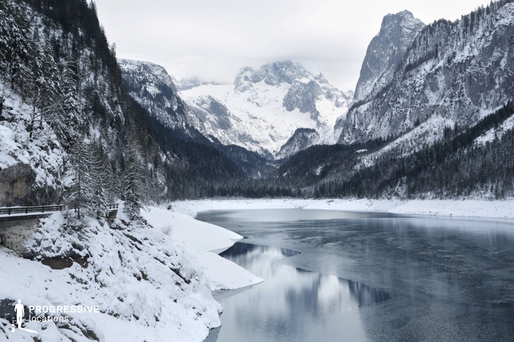 This expansive alpine location features a partially frozen lake nestled between steep, snow-covered mountains. Towering rocky cliffs and densely forested slopes flank the water on both sides, creating a dramatic V-shaped composition that draws the eye toward the rugged, glaciated peaks in the background. The lake’s smooth surface reflects the surrounding landscape, adding symmetry and depth to the scene. Dominated by cool grey, white, and dark green tones, the color palette evokes a stark, serene atmosphere. The terrain is varied, with high elevations, vertical stone faces, and narrow pathways carved into the rock along the lake’s edge. This natural setting, rich in contrast and scale, is ideal for filming genres like fantasy, adventure, or thriller, and supports scenes requiring remote wilderness, majestic panoramas, or isolated, high-altitude environments.