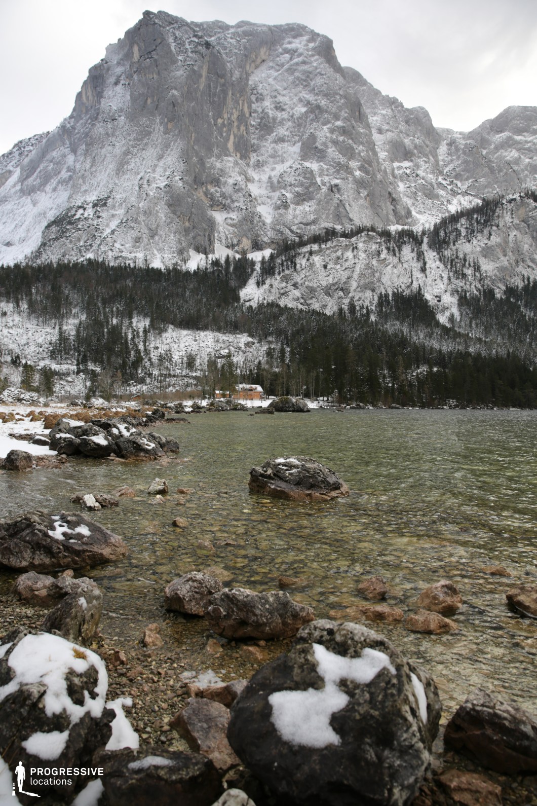 This natural location presents a dramatic lakeside setting dominated by a towering, rugged mountain face with steep vertical rock formations. The foreground features a rocky shoreline with partially submerged stones of various sizes, set in clear, shallow water that reveals a pebbled lakebed. Snow lightly dusts many of the rocks, enhancing the textural contrast between stone and water. The dense pine forest at the mountain’s base provides a dark, natural band that separates the vertical stone wall from the water's edge. The towering cliff in the background forms a powerful compositional element, creating a sense of scale and enclosure. The perspective, captured from a low vantage point near the water, emphasizes depth and invites cinematic exploration of the vertical and horizontal layers. This location is ideal for filming nature-based narratives, contemplative scenes, or atmospheric moments in genres such as drama, thriller, or fantasy, where an imposing and immersive wilderness backdrop is essential.