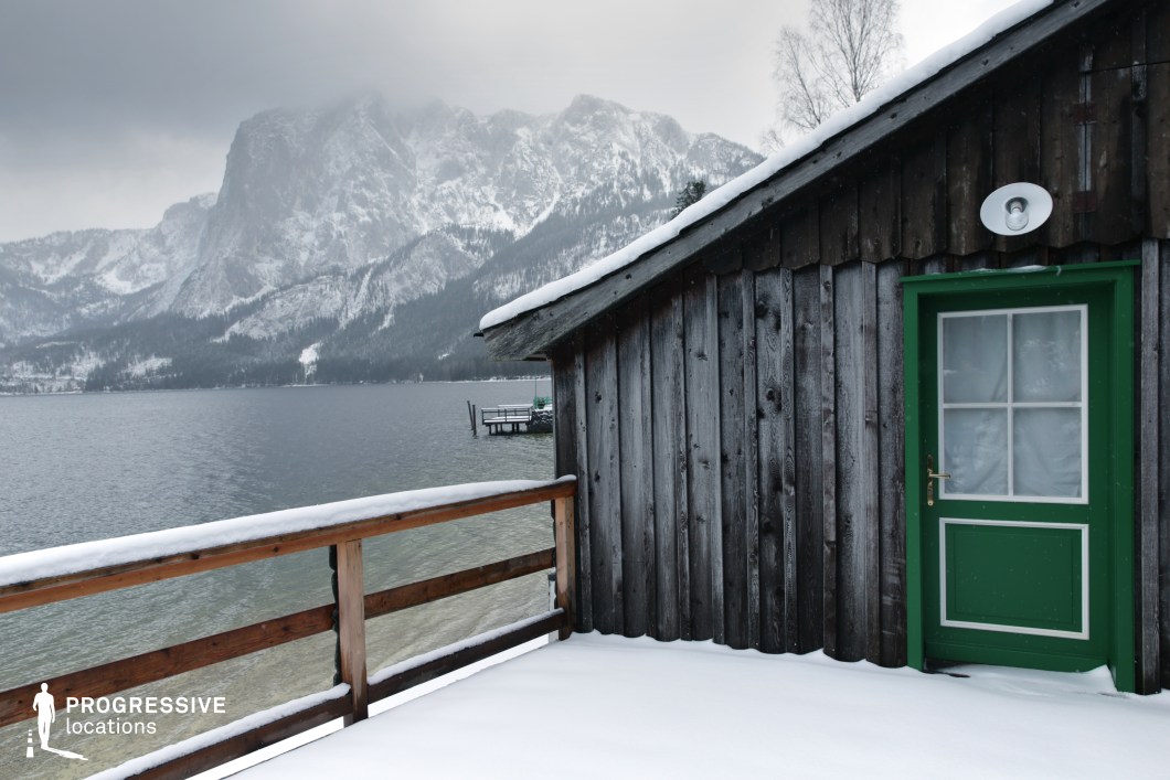 This location features a rustic wooden cabin situated directly beside a calm mountain lake, with a small dock extending into the water and an imposing, snow-covered mountain range in the background. The weathered wood cladding of the structure adds an aged, natural character, while the vivid green door introduces a contrasting color accent that breaks the otherwise monochromatic palette of whites, grays, and natural browns. The simple timber railing frames the scene and reinforces the intimate scale of the lakeside platform. The composition, shot from a slightly low angle, emphasizes the cabin's sturdy form against the vast natural landscape, with strong linear geometry from the roofline and railings guiding the eye into the open space. This location is ideal for filming contemplative, nature-focused scenes or minimalist compositions in drama, arthouse, or thriller genres, offering a serene yet visually rich setting for exterior shots.