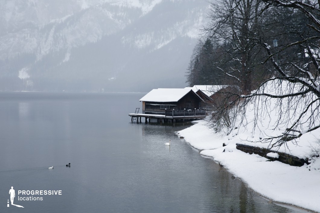This location features a tranquil lakeside scene dominated by a dark wooden cabin on stilts extending over the water, accompanied by a narrow dock and backed by a gently rising, snow-covered hillside. The muted color palette consists of whites, greys, and deep browns, which together create a calm, subdued visual atmosphere. The bare trees with spindly branches add linear structure and contrast against the uniform snow blanket. The water’s still surface merges seamlessly with the soft outlines of the distant, mist-covered mountains, creating a strong sense of spatial depth. This composition is ideal for capturing serene, isolated, or contemplative moods and lends itself well to film genres like drama, mystery, or arthouse cinema. It is especially fitting for scenes requiring quiet natural surroundings or remote, introspective settings.