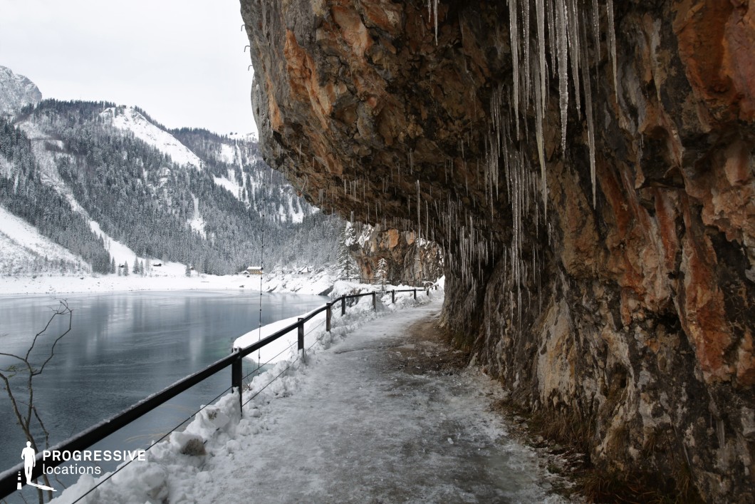 This striking natural location features a narrow, icy path carved into the base of a rugged, overhanging rock formation, which is heavily adorned with long, vertical icicles. The rock face exhibits a rich palette of earthy tones including rust, grey, and brown, with coarse textures that emphasize its raw, untouched character. A simple black railing runs alongside the edge, separating the path from the adjacent frozen lake, which stretches into a backdrop of steep, densely forested mountains. The visual contrast between the solid, jagged cliff wall and the expansive, calm lake surface creates a dramatic depth. The pathway’s confined scale and the looming rock ceiling intensify the sense of enclosure and isolation. This location is ideal for filming suspenseful or introspective scenes, particularly in genres like thriller, survival drama, or mystery, offering a visually immersive and atmospherically charged setting.