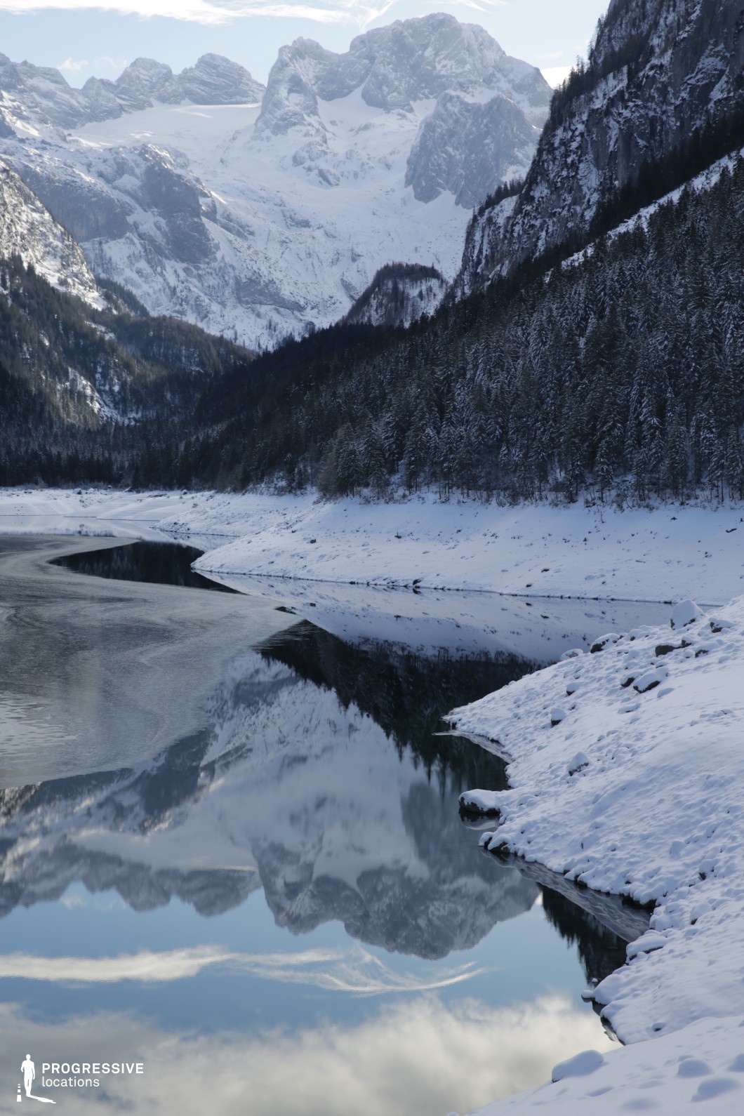 This visually striking location presents a dramatic alpine valley where a still, reflective lake mirrors snow-covered mountain peaks and dense pine forested slopes. The jagged vertical formations of the towering rock faces create a strong contrast with the soft curves of the snow-covered shoreline and forested hills. The composition emphasizes natural symmetry through the lake’s reflection, enhancing the sense of depth and scale. The color palette is dominated by icy whites, deep greys, and cold blues, reinforcing a remote and untouched atmosphere. The vertical framing captures the rising terrain from the foreground snowbank to the towering mountain ridge, providing a powerful sense of vertical continuity. This natural setting is ideal for film productions requiring majestic, isolated, or introspective outdoor environments, suitable for genres such as epic drama, survival, fantasy, or environmental documentary.