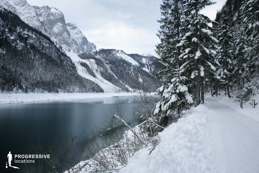 This location features a serene lakeside pathway bordered by dense pine forest, all blanketed in snow, with towering, rugged mountains forming a dramatic backdrop. The lake’s still, dark surface contrasts with the white snow cover, creating a bold visual dichotomy. The towering conifer trees are heavily layered with snow, enhancing the natural symmetry and texture of the composition. The narrow path curves gently, framed by trees on one side and the lake on the other, offering a sense of enclosure yet openness toward the mountainous expanse. The overall visual structure is defined by clean horizontal and vertical lines formed by the shoreline, the tree trunks, and the mountain ridges. This setting is ideal for productions seeking a pristine, remote, and contemplative natural environment. It lends itself well to genres such as drama, thriller, or fantasy, and is suited for scenes involving isolation, transition, or introspection in a visually immersive landscape.