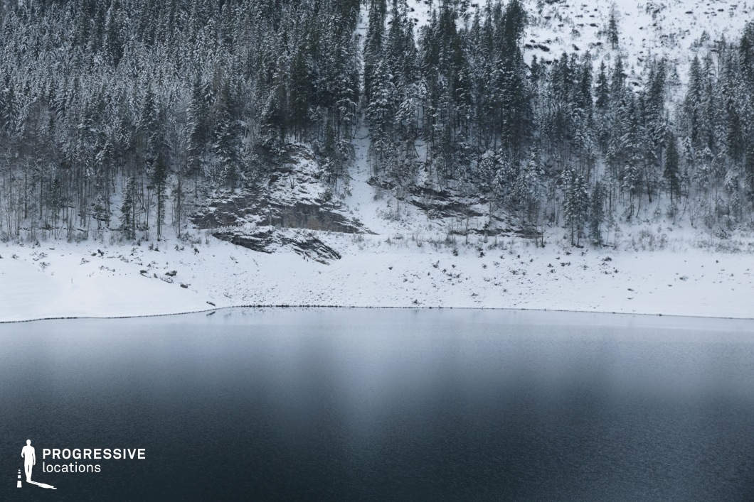 This location features a serene, snow-blanketed forested hillside descending into a still, dark lake. The slope is densely populated with tall, coniferous trees uniformly dusted with snow, creating a visually cohesive and texturally rich backdrop. In the midsection of the slope, exposed layered rock formations emerge from beneath the forest, introducing a rugged, natural element that contrasts with the soft texture of the surrounding snow. The water in the foreground is smooth and mirror-like, offering a stark horizontal contrast to the vertical rhythm of the tree trunks. The natural curve of the shoreline adds depth and guides the eye laterally across the composition. The scene exudes tranquility and isolation, making it particularly well-suited for filming atmospheric, contemplative, or suspenseful sequences. Its untouched, remote aesthetic is ideal for nature documentaries, dramas, or psychological thrillers, and it provides a versatile canvas for wide landscape shots or character-centric scenes within a powerful natural setting.