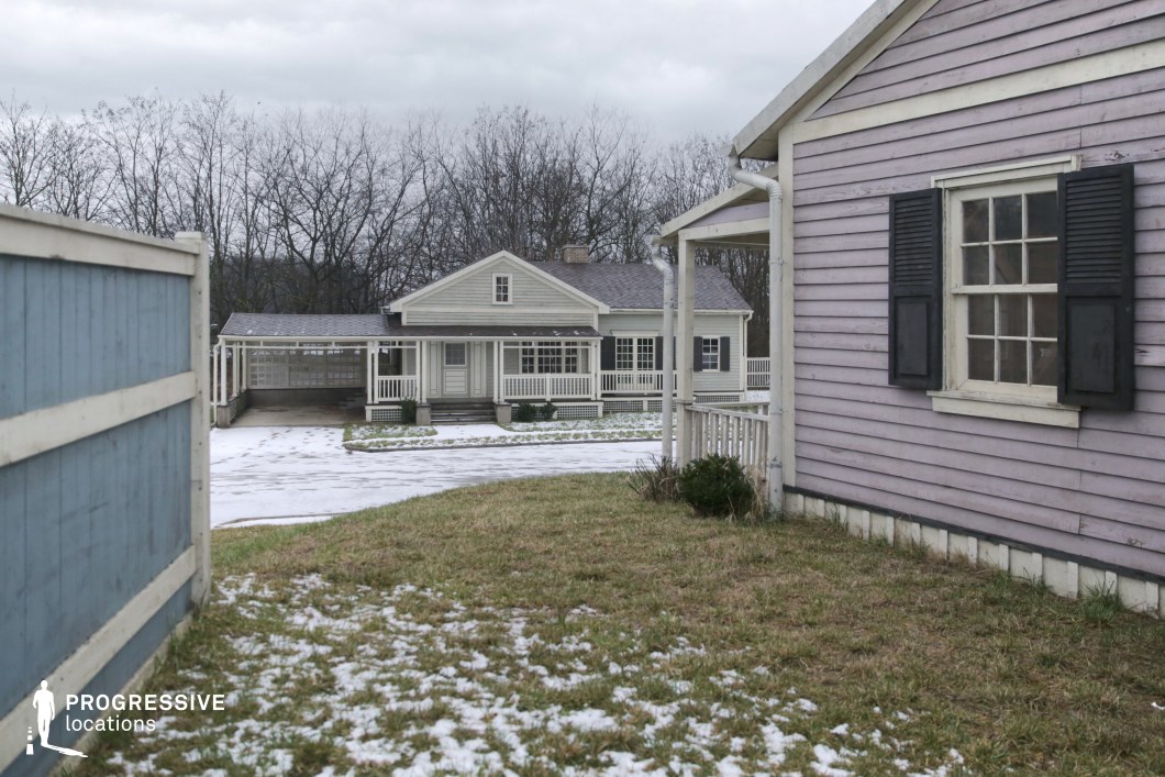 This location presents a well-maintained residential setting defined by wooden siding structures painted in muted pastel tones. The lavender-colored building in the foreground features classic window shutters and horizontal plank cladding, adding a rustic charm. The background reveals a symmetrical, light-grey house with a full-width porch, vertical railings, and an attached carport, all contributing to a cohesive American suburban style. The open grassy yard with sparse snow coverage enhances the sense of seasonal transition without dominating the composition. The visual framing between the two houses and the fencing creates a natural corridor, adding depth and layered perspective. Ideal for filming intimate domestic narratives, suburban dramas, or retro-period pieces, the scene offers an authentic and versatile backdrop.