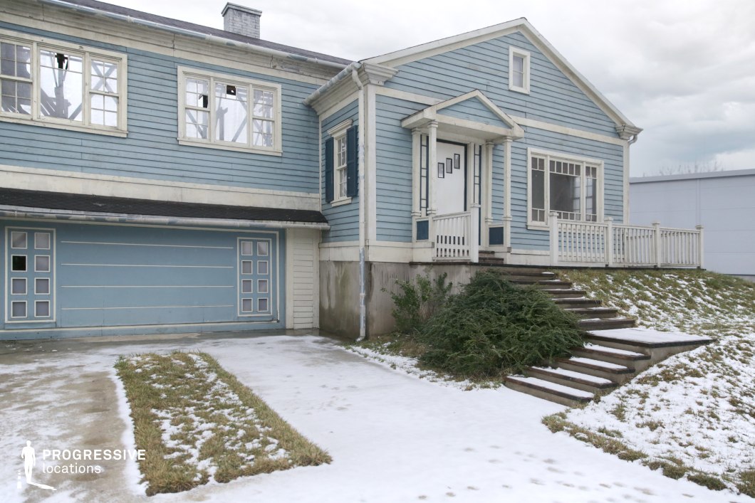 This location features a light blue, wooden-clad suburban-style house with visible signs of wear and age, including chipped paint, stained concrete, and broken upper windows. The structure includes a prominent double garage with geometric window patterns, a slightly raised entrance with classic white columns and railing, and a wide front staircase integrated into a sloped, partially snow-covered lawn. The symmetry and repetition of rectangular elements across doors, windows, and siding create a visually organized yet subtly unsettling atmosphere. The muted color palette and structural degradation lend the scene a sense of abandonment or artificiality, making it particularly suitable for filming psychological thrillers, dystopian or horror genres, and scenes requiring a stylized, uncanny suburban backdrop.