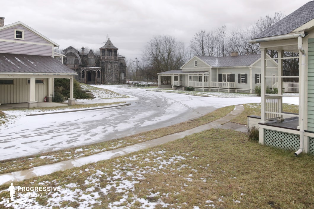 This residential streetscape blends traditional wooden-frame suburban homes with front porches and pastel siding, contrasted starkly by a striking, dark gothic-style mansion positioned at the end of the curved road. The symmetrical façades of the lighter houses contribute to a quaint, orderly atmosphere, while the intricate, decaying architecture of the mansion introduces a dramatic focal point with strong vertical and textural elements. The paved street and front lawns create a spacious, navigable foreground, enhancing depth and leading the viewer’s eye toward the central architectural contrast. This location offers a visually rich setting ideal for film genres such as horror, psychological thrillers, or suburban dramas, providing distinct mood variations within a single frame and enabling diverse interior-exterior transition shots.