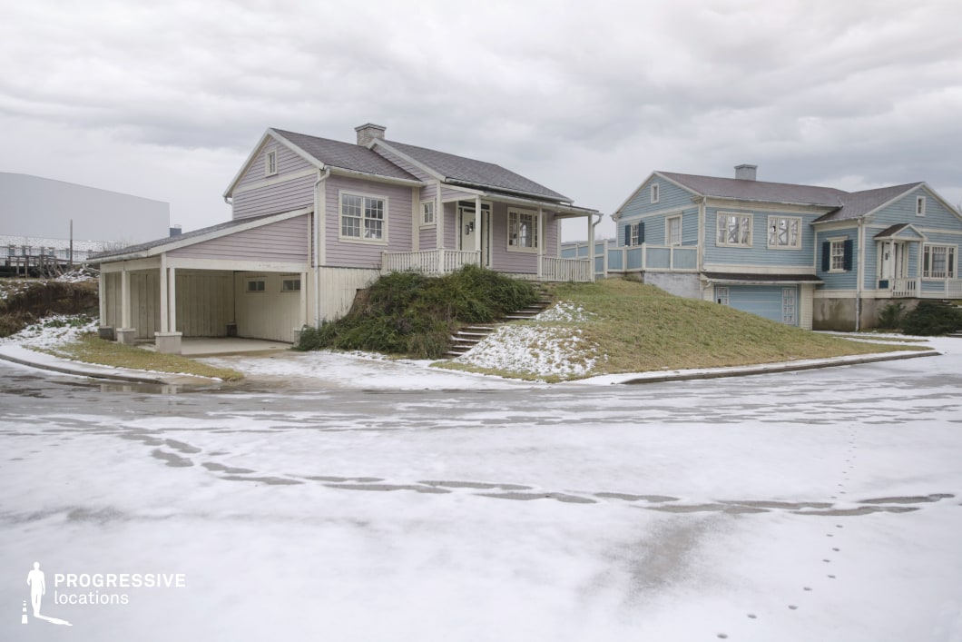 This location presents a pair of pastel-colored, wooden-clad suburban houses with pitched roofs, front porches, and built-in garages, arranged on slightly elevated lawns beside a curved residential road. The stylized architecture, clean lines, and uniform façades give the scene a staged or artificial quality, further emphasized by the absence of surrounding context or detailing. The surface of the road is partially covered in snow, highlighting the structured geometry of the neighborhood layout. The setting evokes a sense of stillness and spatial symmetry, making it particularly effective for shooting stylized suburban narratives, psychological thrillers, or retro-inspired dramas where an idealized or uncanny small-town backdrop is required.