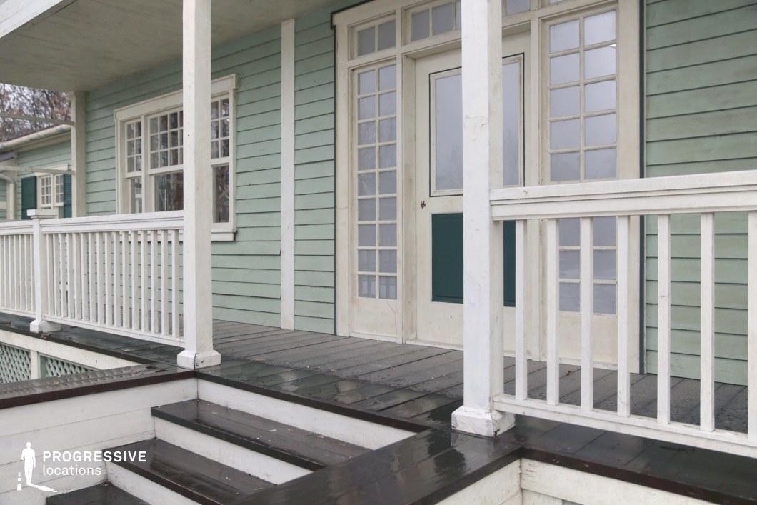 This location presents a quaint, wooden porch attached to a building clad in pastel green horizontal siding, accented by white trim and railings. The symmetrical window panels and central door, both featuring multiple glass panes in a grid layout, lend a balanced and charming architectural character. The dark-stained wooden steps and floorboards create a subtle contrast against the lighter structural elements, while the vertical posts and railing slats reinforce a clean, orderly geometry. The overall aesthetic evokes a serene, small-town or coastal atmosphere with vintage undertones. This setting is ideal for filming period dramas, romantic stories, or suburban-themed productions, particularly for entrance scenes, reflective moments, or character introductions.