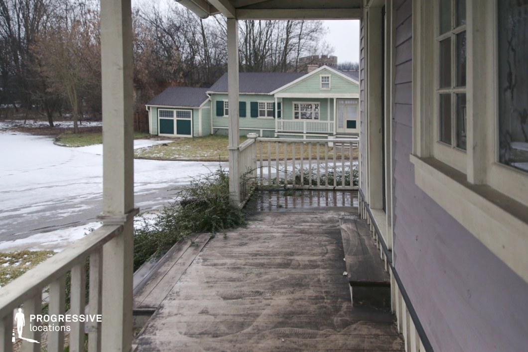 This location features a classic wooden porch with railings and vertical supports, opening onto a quiet residential cul-de-sac lined with single-story, pastel-toned wooden houses. The foreground structure has muted lavender siding and cream-colored trim, with visible wear on the porch floorboards that adds an authentic, lived-in quality. The adjacent mint-green house, complete with shuttered windows, a front-facing gable, and an integrated garage, reinforces the mid-20th-century suburban aesthetic. Surrounded by leafless trees and modest landscaping, the setting creates a sense of openness and calm. This versatile suburban backdrop is well-suited for drama, coming-of-age films, or period productions requiring a small-town American residential environment.