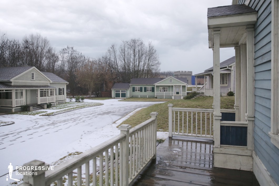 This location showcases a neatly arranged suburban cul-de-sac lined with pastel-colored, single-story wooden houses, each featuring prominent front porches with railings and steps. The architecture is consistent, evoking a mid-20th century American small-town aesthetic, with gabled roofs, shuttered windows, and ornamental trim. The road curves gently through the neighborhood, creating a sense of depth and spatial layering, while the houses are spaced to allow clear sightlines and multiple camera angles. The open foreground with the porch railing adds a natural frame to the composition. This setting is ideal for filming residential or domestic scenes and lends itself well to family dramas, period pieces, or psychological thrillers that require a quiet, controlled suburban environment with classic Americana undertones.