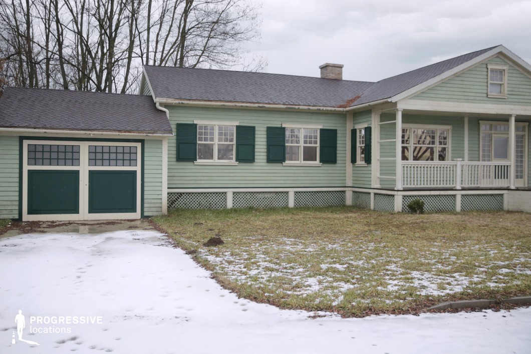 This single-story wooden house showcases classic suburban architecture with a light mint-green facade, dark green shutters, and a gabled roof with asphalt shingles. The horizontal clapboard siding, modest front porch with white balustrades, and symmetrical window layout contribute to its quaint, orderly appearance. A two-door garage on the left side integrates seamlessly with the home’s design, while the lattice skirt detail at the base enhances the cottage-like charm. The partially snow-covered lawn and bare trees in the background emphasize the structure’s clean lines and isolated presence. This location is well-suited for filming suburban residential scenes, particularly for genres such as drama, mystery, or coming-of-age stories, where a quiet, small-town setting is essential.