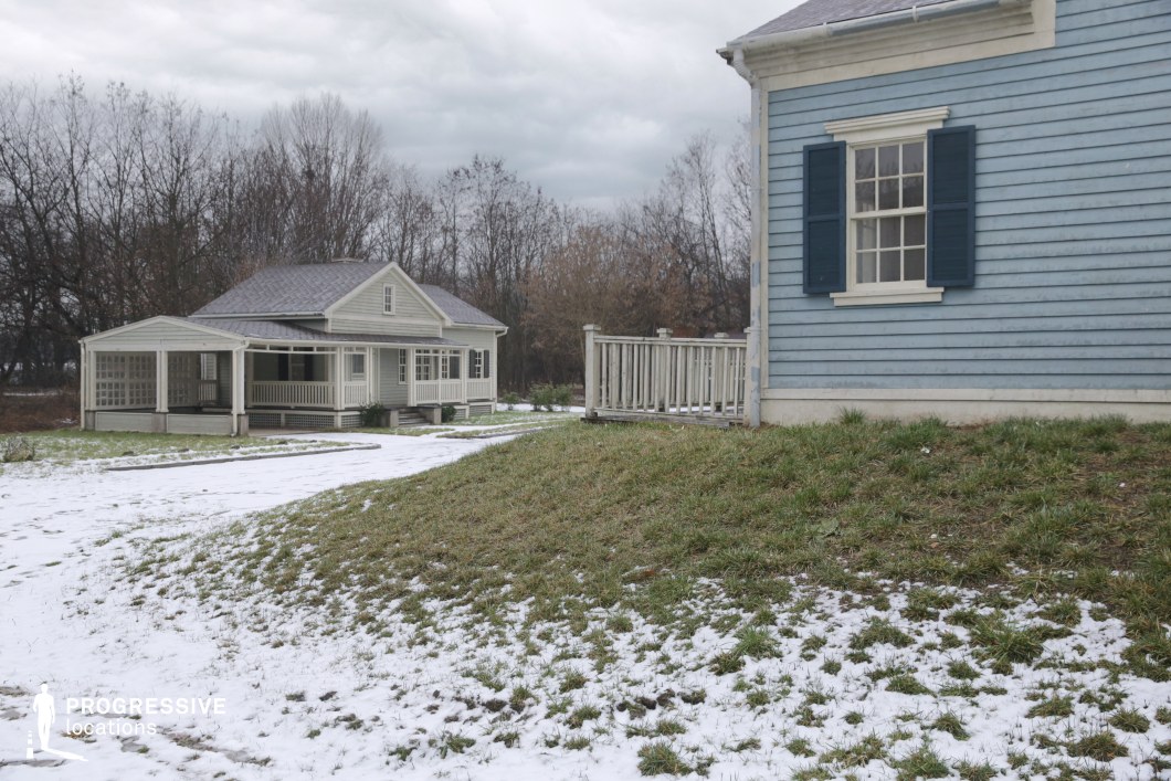 This rural residential setting features two traditional wooden houses with horizontal siding, white-painted railings, and classic shuttered windows. The structures follow a modest, symmetrical architectural style reminiscent of early American vernacular homes, with gable roofs and raised porches that contribute to a homely, timeless character. The pale beige and muted blue tones create a subdued, neutral palette that blends naturally with the wintery ground and bare tree backdrop. The open green area with a gentle incline in the foreground adds spatial depth, enhancing visual layering. This quiet, well-preserved setting is well-suited for period dramas, introspective narratives, or rural lifestyle features, offering a peaceful and authentic location for emotionally focused or dialogue-driven scenes.