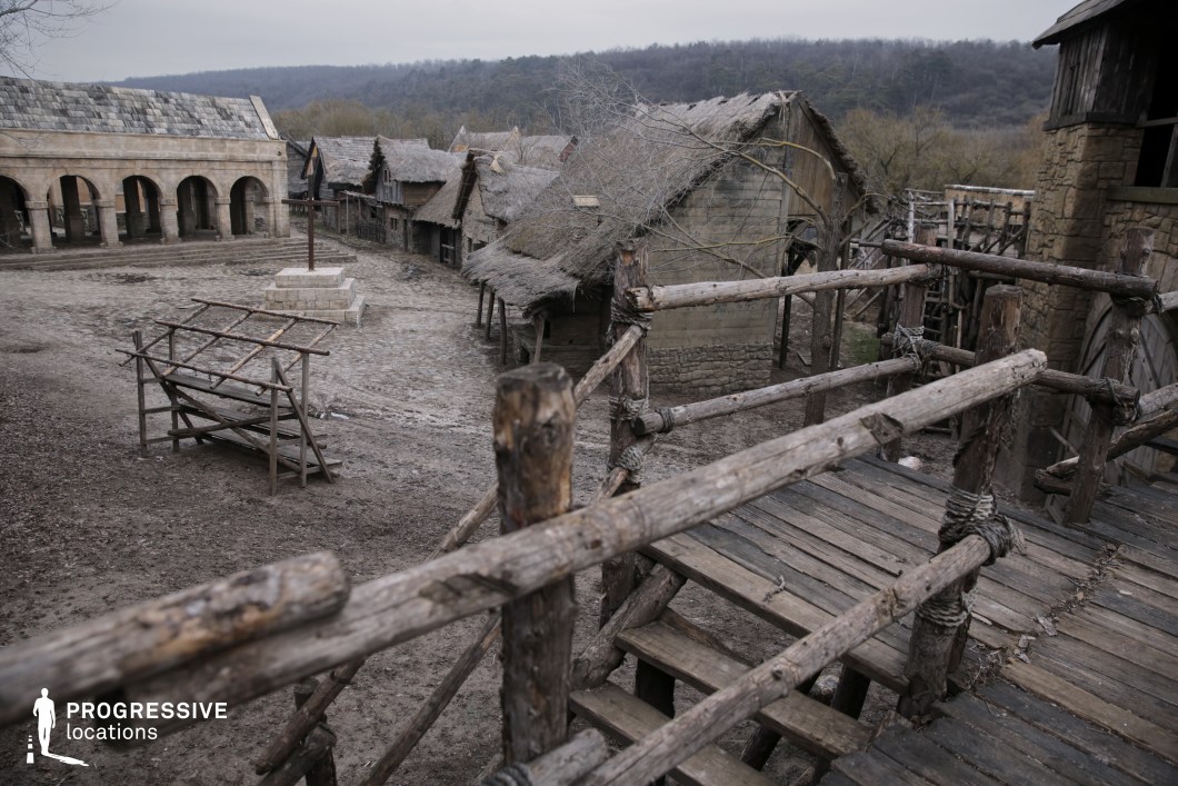 Captured from a rustic wooden walkway, this scene reveals a weathered medieval village square framed by thatched-roof huts and a stone hall with arched arcades. The rough-hewn timber structures, exposed framework, and uneven terrain add a raw, immersive realism. The open plaza space with a raised central platform enhances the setting’s dramatic potential. This location is perfect for fantasy, historical, or medieval drama productions, ideal for scenes involving public speeches, executions, village life, or battles erupting in a central square.