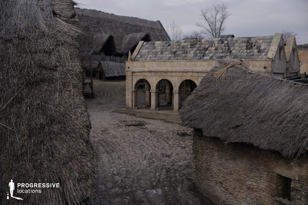 This rustic settlement features a blend of primitive stone buildings and thatched-roof huts, connected by muddy, uneven streets. A central structure with arched stone colonnades introduces an element of authority or communal gathering, contrasting with the handmade, organic appearance of the surrounding dwellings. The scene evokes a sense of pre-modern or early medieval life, with authentic textures and a natural layout ideal for immersive storytelling. This location is particularly well-suited for historical dramas, tribal or peasant life depictions, fantasy worlds, or scenes set in ancient or early medieval villages.