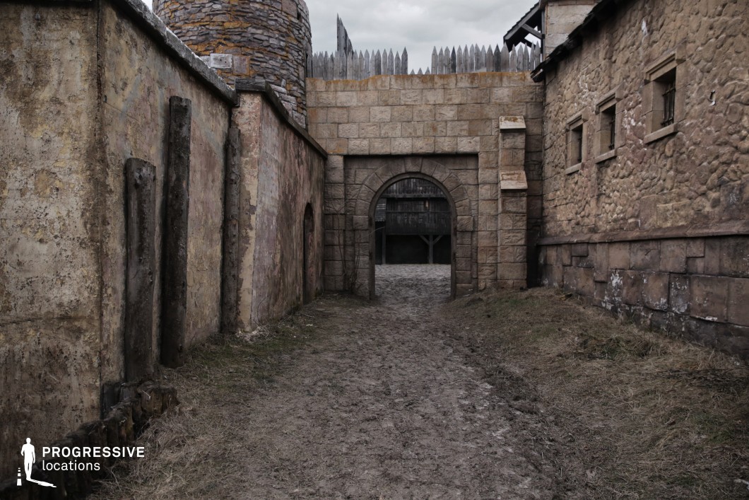 This enclosed passage leads through a robust arched stone gate, flanked by coarse plaster and cobblestone buildings, and backed by palisade fencing and a rounded turret. The mix of rough textures, aged surfaces, and compact design evokes the entrance to a fortified town or castle interior. The narrow layout and layered depth offer excellent framing opportunities for suspenseful or dramatic entrances. This location is ideal for medieval, fantasy, or historical productions, especially scenes involving patrols, secret escapes, guarded transitions between town sectors, or looming threats at the gates.