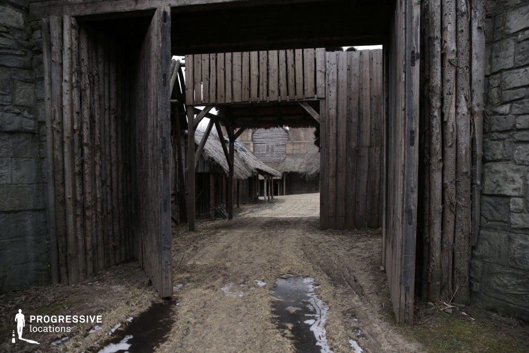 This setting captures a rugged wooden gate embedded in a stone wall, leading into a primitive settlement lined with timber structures and thatched roofs. The layers of fencing, gates, and overhanging wooden watch platforms create a fortified, defensive atmosphere typical of early medieval or tribal settlements. The muddy, uneven pathway adds realism and texture to the scene. This location is ideal for historical epics, medieval fantasies, or tribal village stories, particularly for establishing shots of a guarded entrance, siege preparations, or scenes involving arrivals and departures from a fortified outpost.