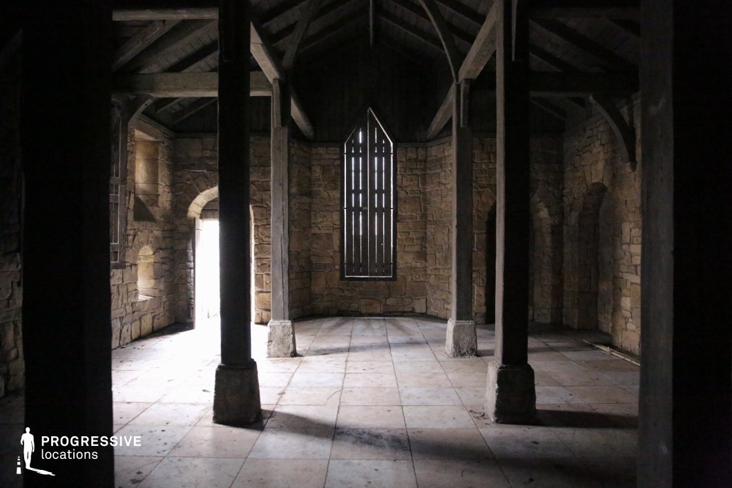 This space reveals a dimly lit, vaulted chamber with exposed timber beams, thick stone walls, and a narrow arched window, evoking the austerity of a small medieval chapel or monastic hall. The symmetrical pillars and modest floor tiles contribute to a solemn, timeless atmosphere, suggesting reflection, secrecy, or ancient tradition. Ideal for period dramas, fantasy series, or mystery films, this location is well-suited for scenes involving sacred rituals, secret gatherings, solitary prayer, or quiet moments of revelation within a spiritual or historical setting.