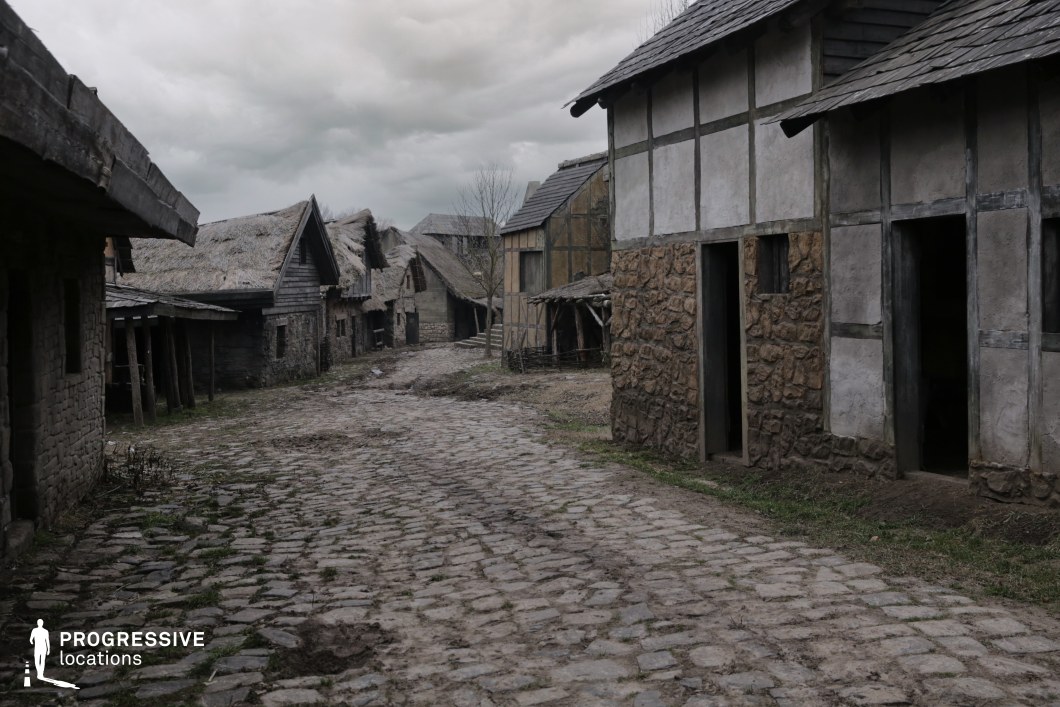 This scene features a winding cobblestone street lined with weathered, half-timbered houses and thatched or shingled rooftops, evoking a modest rural settlement from the Middle Ages. The rough textures, asymmetrical building lines, and earthy palette give the location a grounded, lived-in quality, suggesting simplicity, hardship, or authenticity. This setting is ideal for filming historical dramas, medieval fantasies, folklore-based stories, or scenes portraying everyday life, village unrest, or encounters between travelers and locals in a pre-modern world.