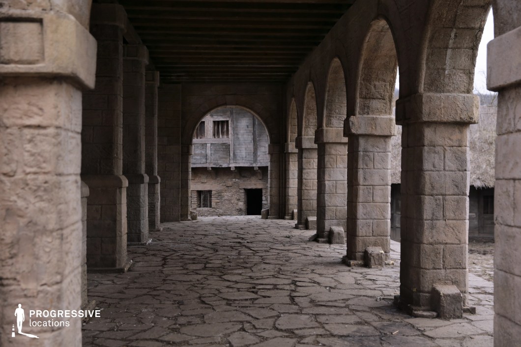This image features a long, vaulted stone arcade with evenly spaced rectangular columns supporting rounded arches, all constructed from uniformly textured blocks. The cobblestone floor enhances the rustic and aged appearance of the walkway, which leads toward a dimly lit structure composed of weathered stone and wood. The linear arrangement of the arches creates a strong geometric rhythm and depth, ideal for leading the viewer’s eye into the composition. The combination of architectural symmetry and natural erosion gives the space an immersive, historical character. This location is highly suited for period dramas, medieval productions, or fantasy narratives, offering a versatile setting for suspenseful transitions, processions, or atmospheric dialogue scenes.